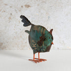 A small metal bird sculpture with a distressed teal, brown, and black patina body, orange legs, and a dark tail.