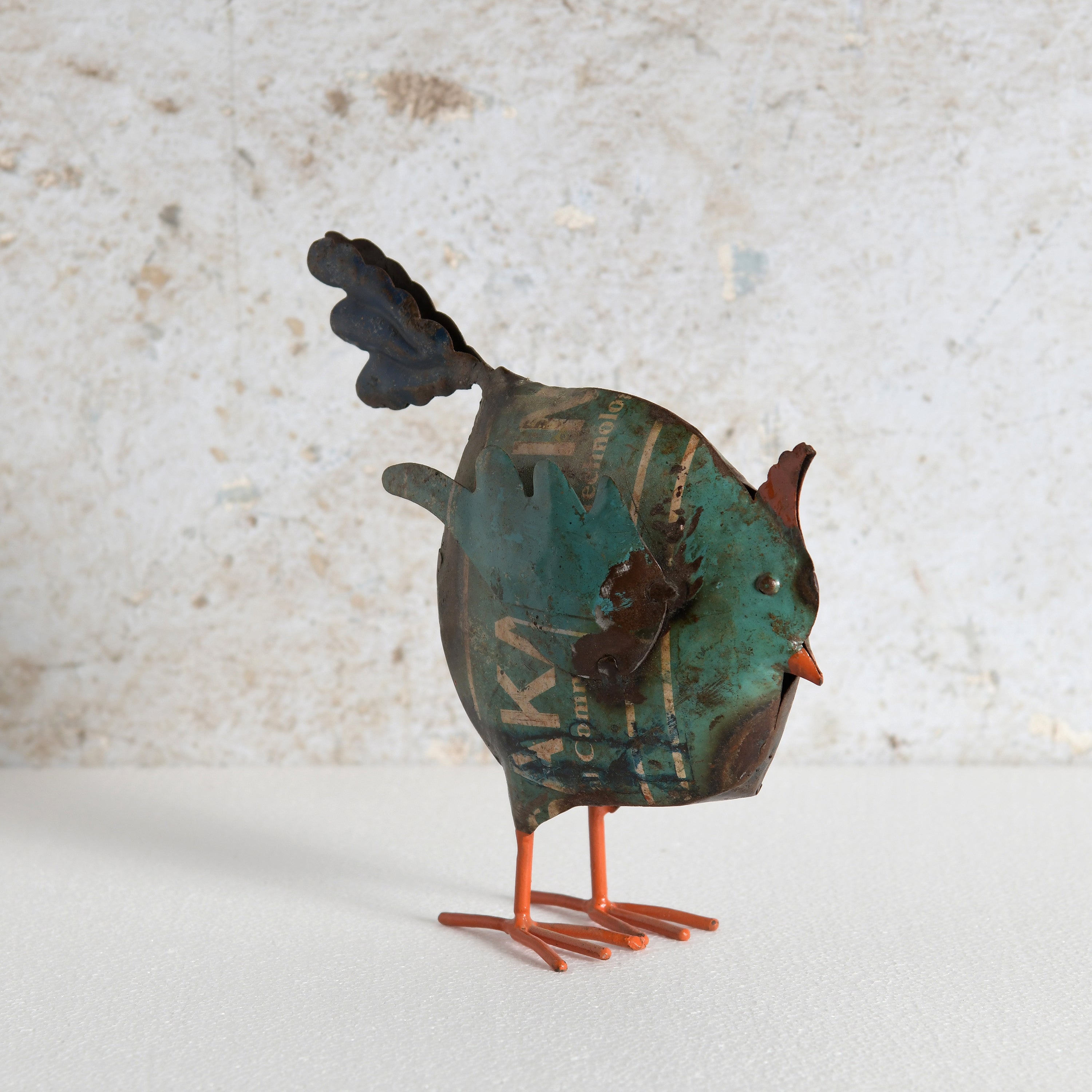 A small metal bird sculpture with a distressed teal, brown, and black patina body, orange legs, and a dark tail.