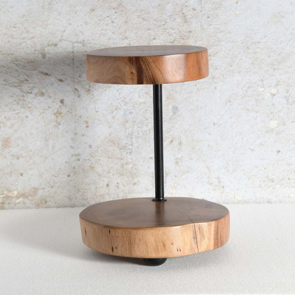 A modern two-tier wooden platter stand featuring circular wood slices at the top and bottom tiers connected by a slim black metal pole.