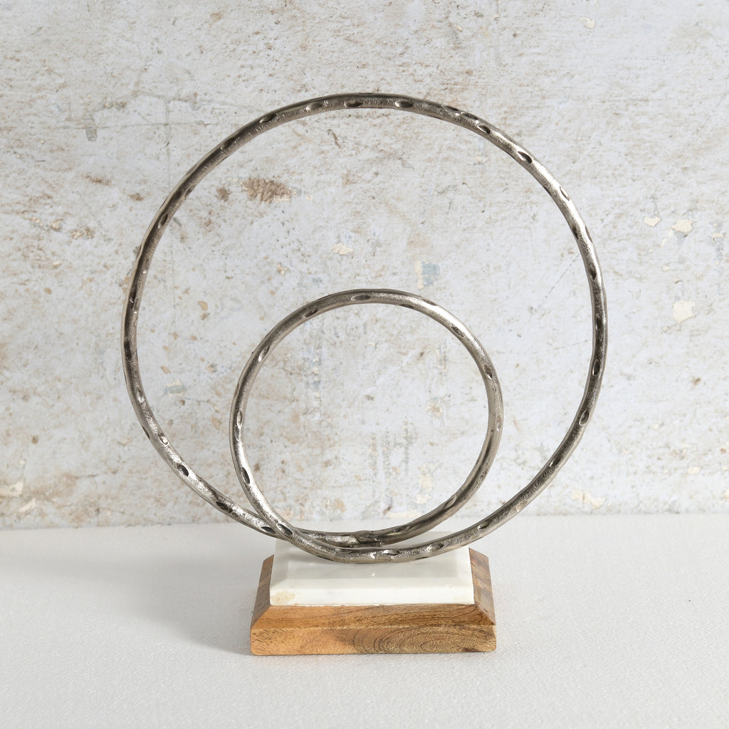 A decorative metal sculpture featuring two interlocking hammered circular rings mounted on a rectangular wooden base with a white inlay.