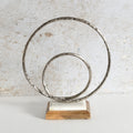 A decorative metal sculpture featuring two interlocking hammered circular rings mounted on a rectangular wooden base with a white inlay.