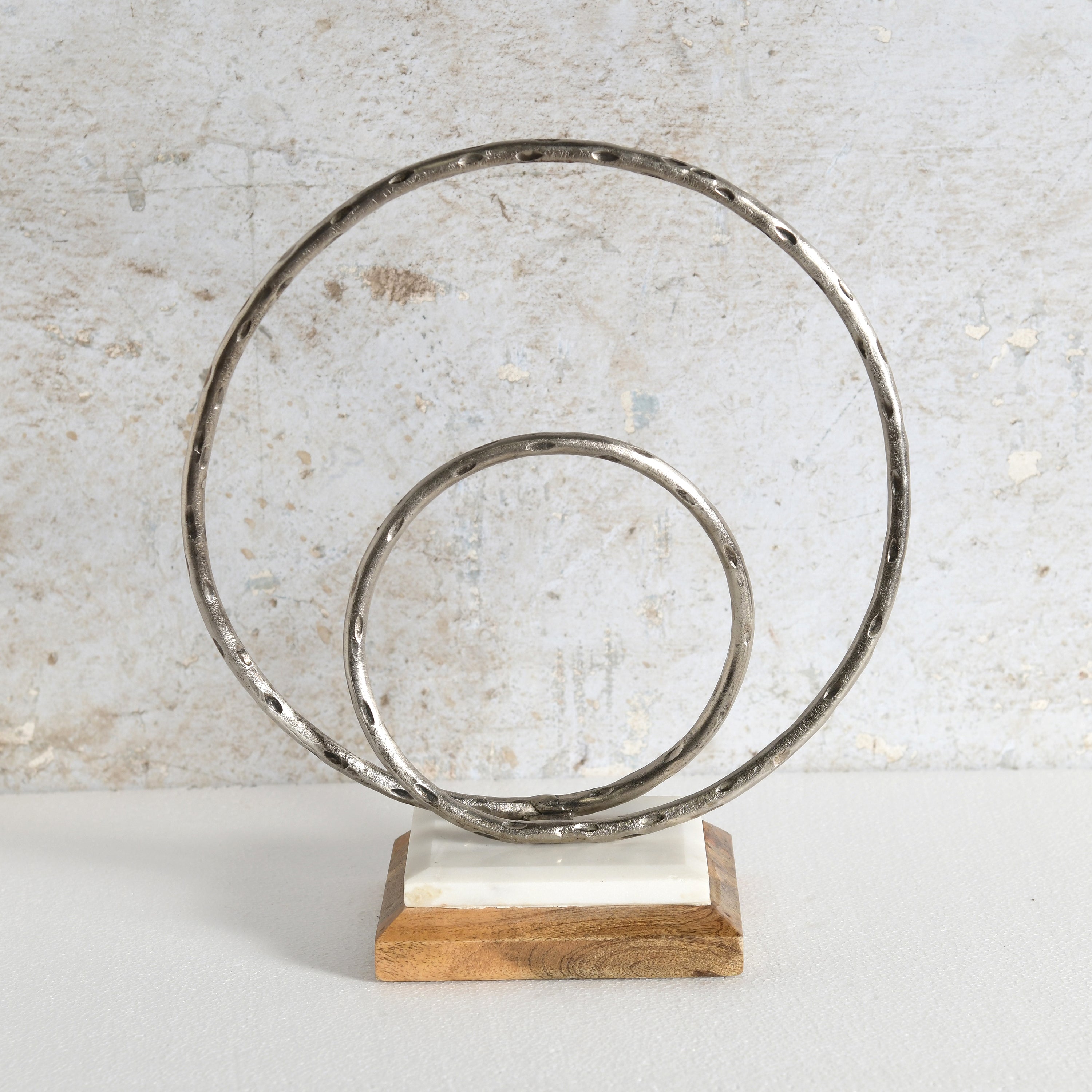 A decorative metal sculpture featuring two interlocking hammered circular rings mounted on a rectangular wooden base with a white inlay.