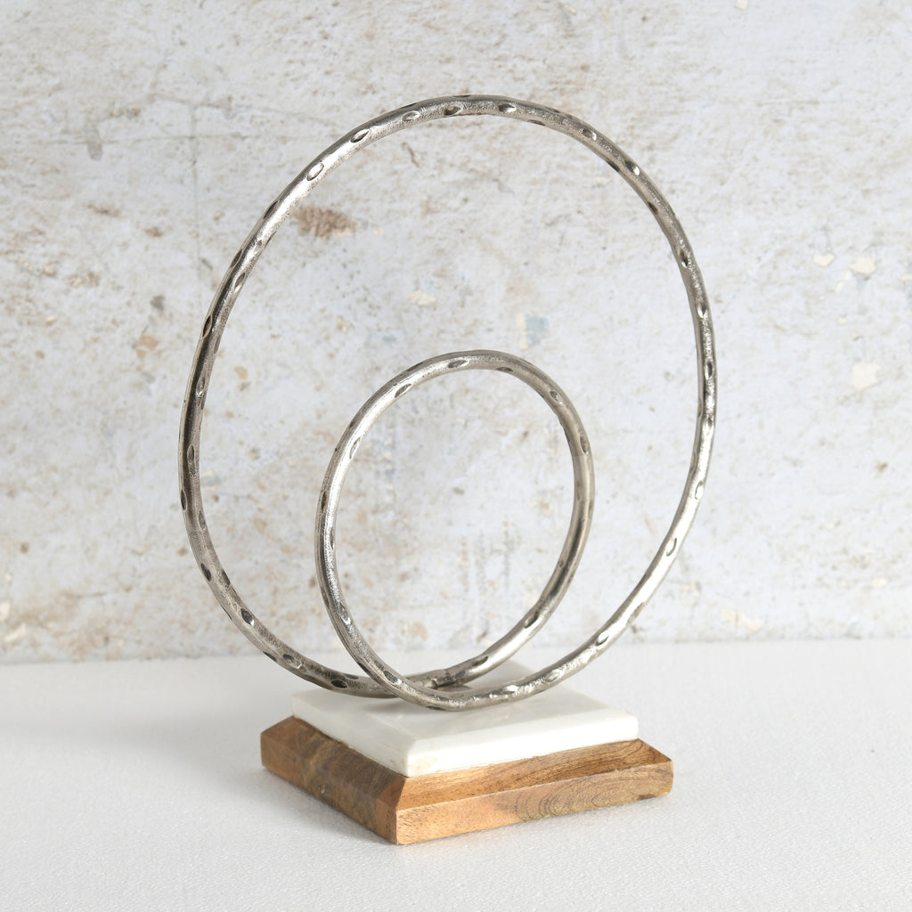 A decorative abstract sculpture with two hammered metal rings in a spiral arrangement, mounted on a white stone slab and a natural wood base.