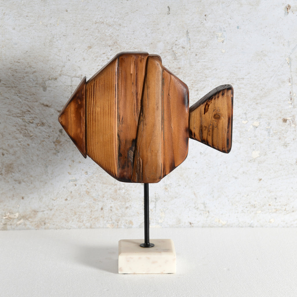 A decorative wooden fish sculpture with a warm medium-brown finish and visible wood grain, mounted on a thin black stand over a small white marble base.