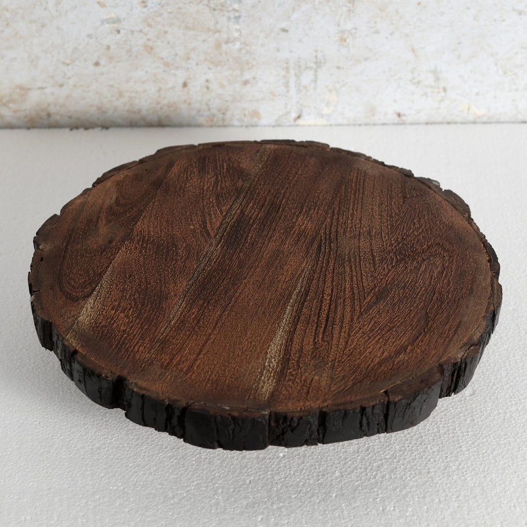 A round wooden serving board or tray | Lazy Susan made from a natural tree slice, featuring a rich dark brown wood grain on top and a dark, rough bark edge, set against a light textured background.