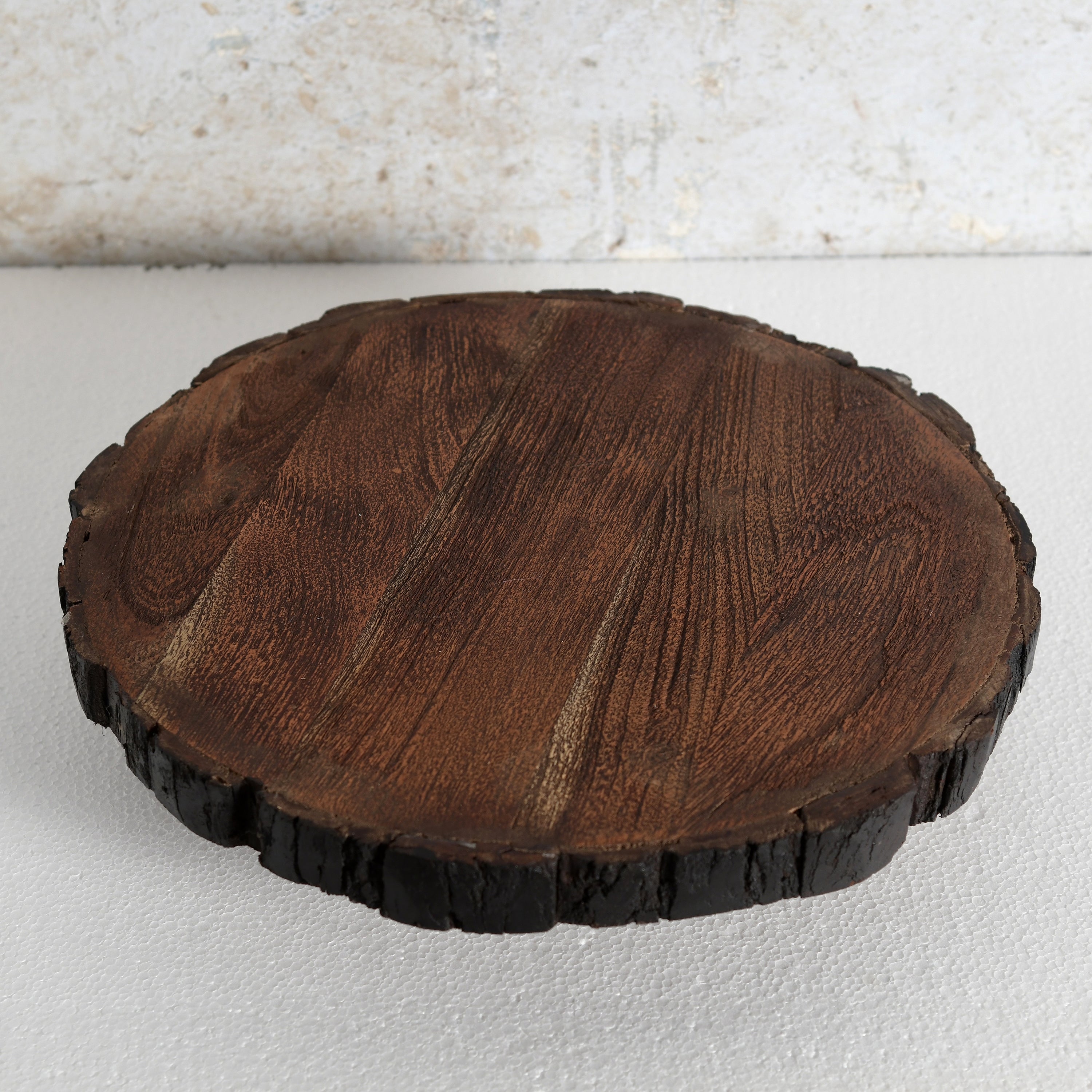A round wooden serving board or tray | Lazy Susan made from a natural tree slice, featuring a rich dark brown wood grain on top and a dark, rough bark edge, set against a light textured background.