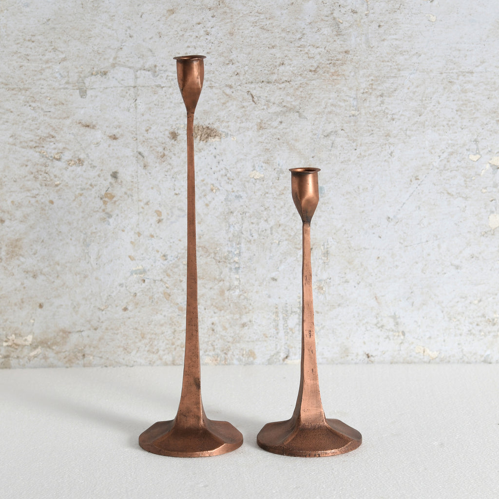 Two vintage copper candlesticks of different heights standing on a light surface against a textured off-white wall.