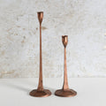 Two vintage copper candlesticks of different heights standing on a light surface against a textured off-white wall.