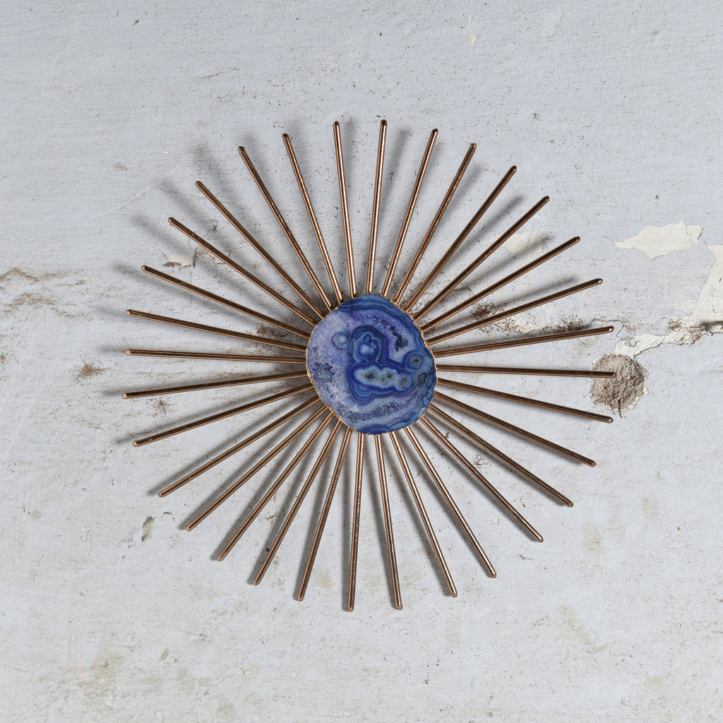 A vintage-style sunburst wall mirror with bronze-toned metal rays and a central blue agate cabochon, mounted on a distressed off-white plaster wall.