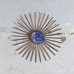 A vintage-style sunburst wall mirror with bronze-toned metal rays and a central blue agate cabochon, mounted on a distressed off-white plaster wall.