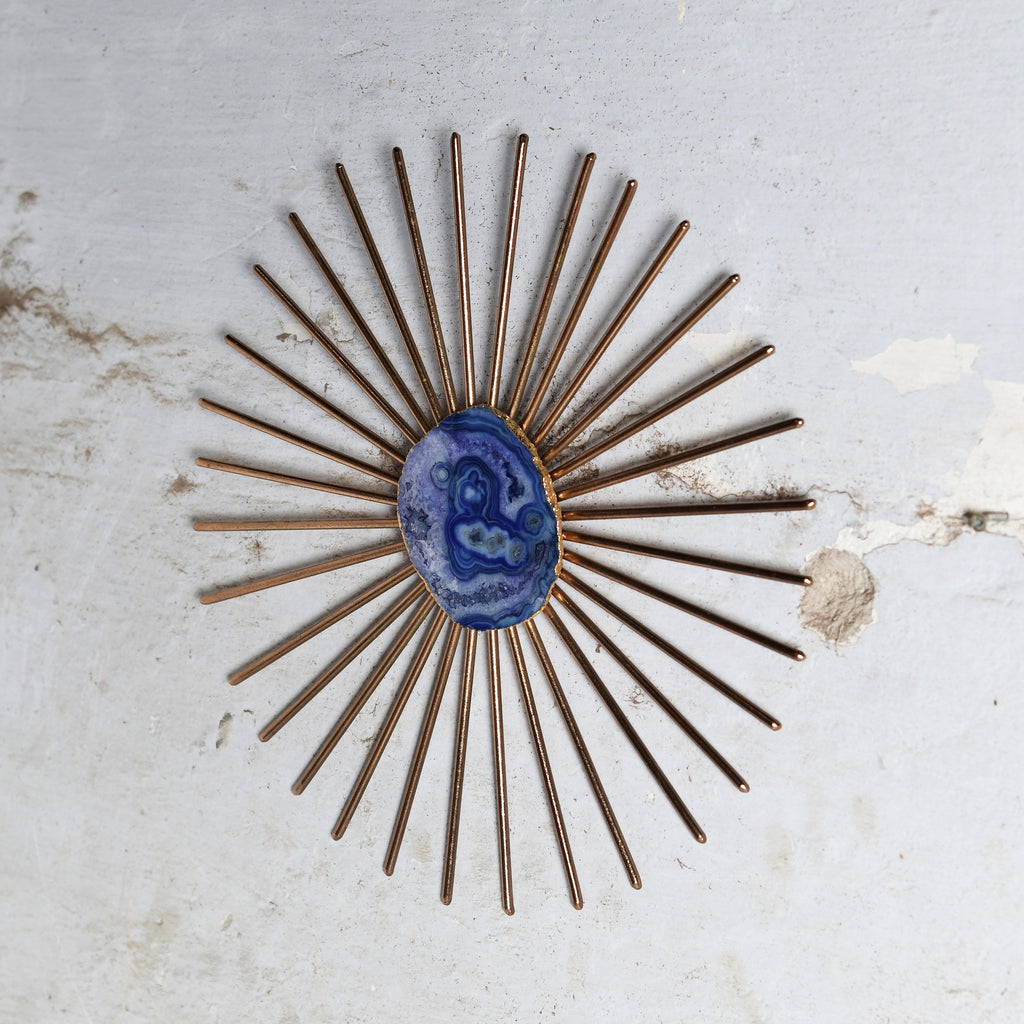 A vintage-style sunburst wall decor with bronze-colored metal rays and a central oval blue agate stone, displayed on a distressed off-white textured wall.
