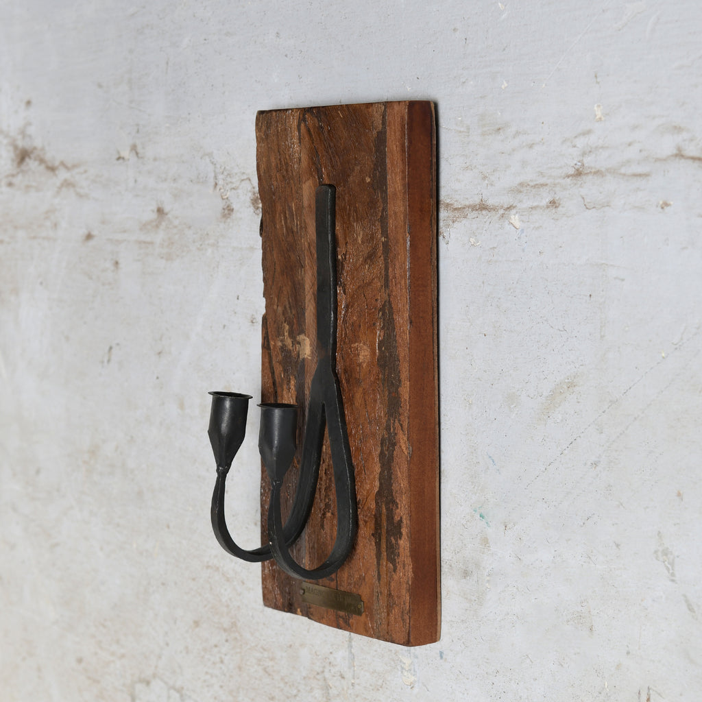 A rustic wall-mounted candle holder featuring a rectangular brown wooden plaque with two black metal candle holder.