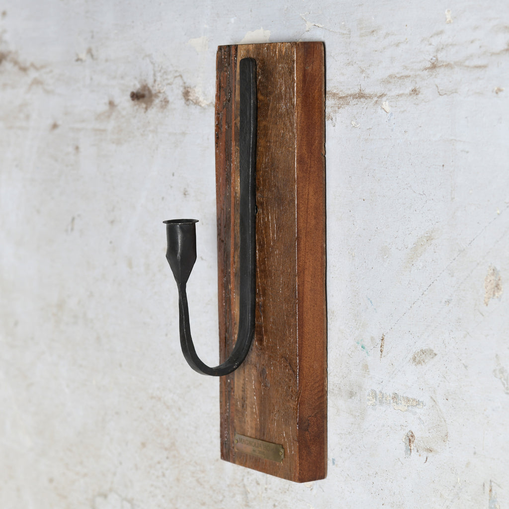 A rustic wall-mounted candle holder featuring a curved black metal candle sleeve on a rectangular brown wooden plaque.