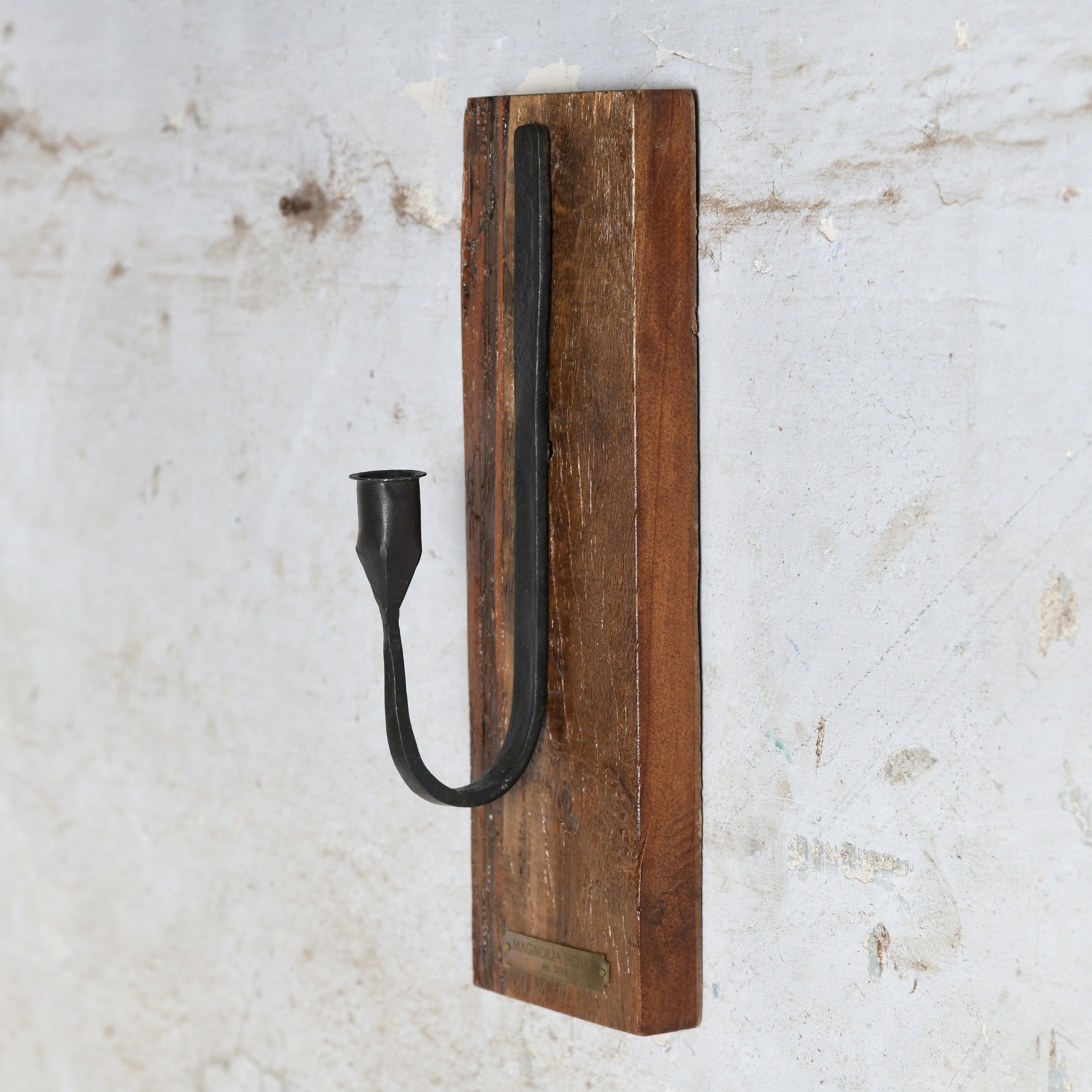 A rustic wall-mounted candle holder featuring a curved black metal candle sleeve on a rectangular brown wooden plaque.