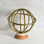 A decorative brass globe sculpture with a spherical wireframe design, mounted on a white circular base and a natural wood pedestal.