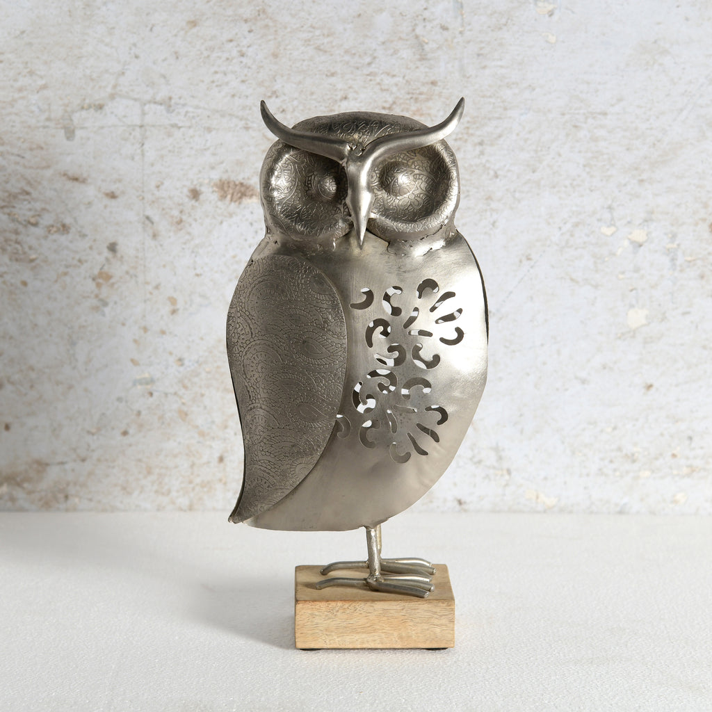 A metal owl figurine with a textured silver finish and decorative cutouts on its body, standing on a small rectangular wooden base against a light textured wall.