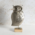 A metal owl figurine with a textured silver finish and decorative cutouts on its body, standing on a small rectangular wooden base against a light textured wall.