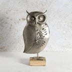 A metal owl figurine with a textured silver finish and decorative cutouts on its body, standing on a small rectangular wooden base against a light textured wall.