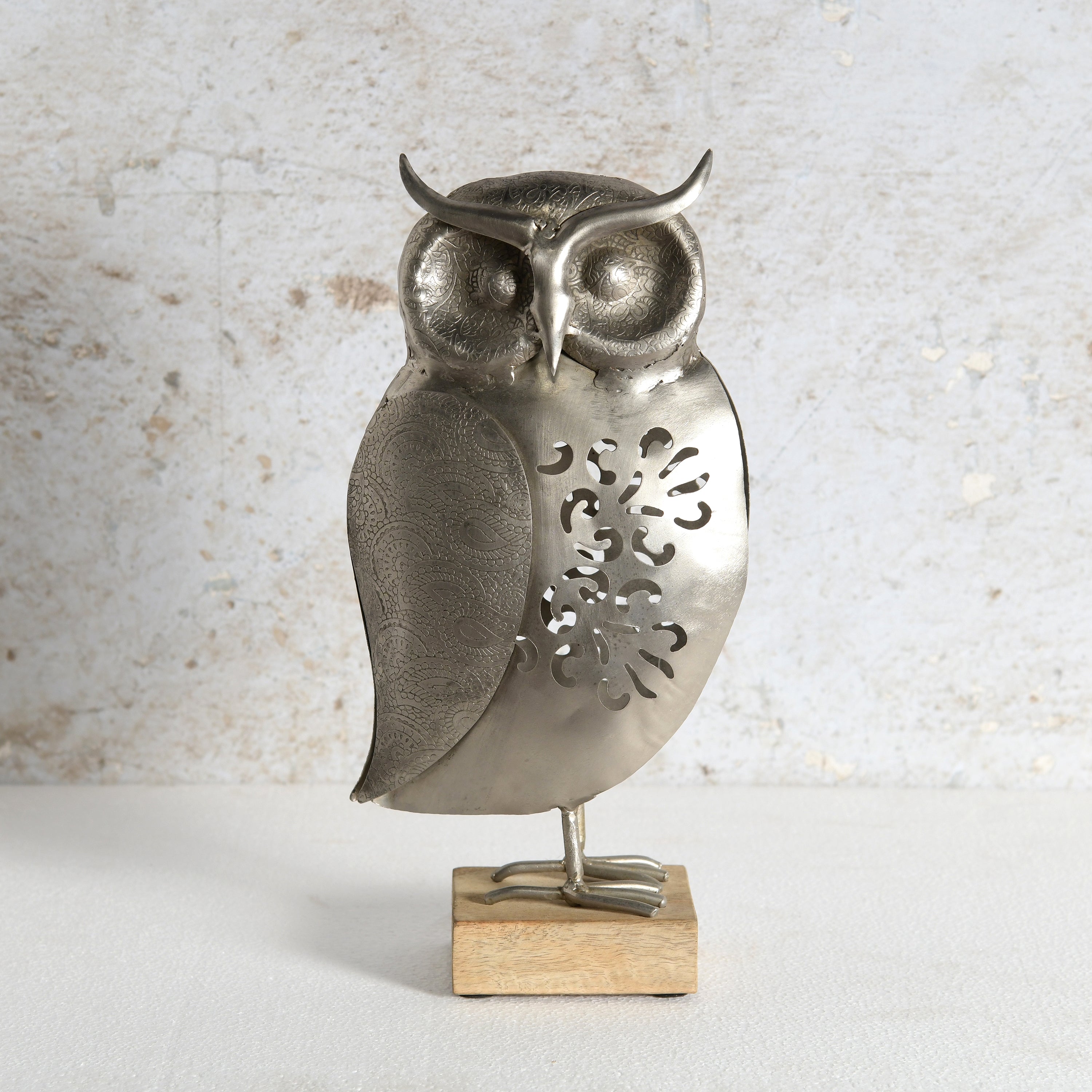 A metal owl figurine with a textured silver finish and decorative cutouts on its body, standing on a small rectangular wooden base against a light textured wall.