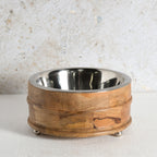 A round pet feeder with a polished stainless steel interior and a carved wooden exterior, resting on small metal feet.